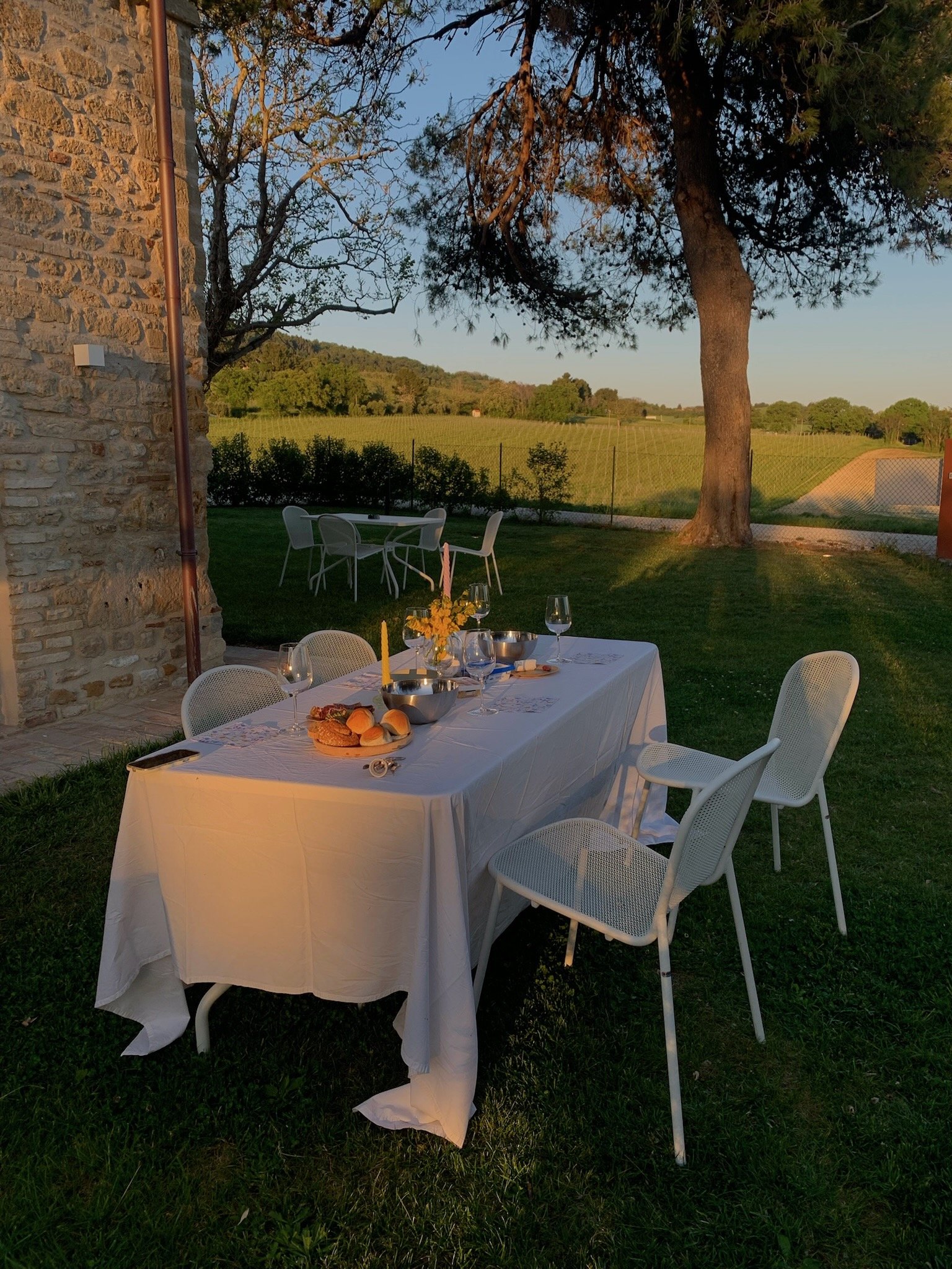 Tavolo apparecchiato all'aperto con tovaglia bianca, piatti, bicchieri, pane e candele, situato in giardino con vista su campagna e alberi, illuminato dalla luce del tramonto.