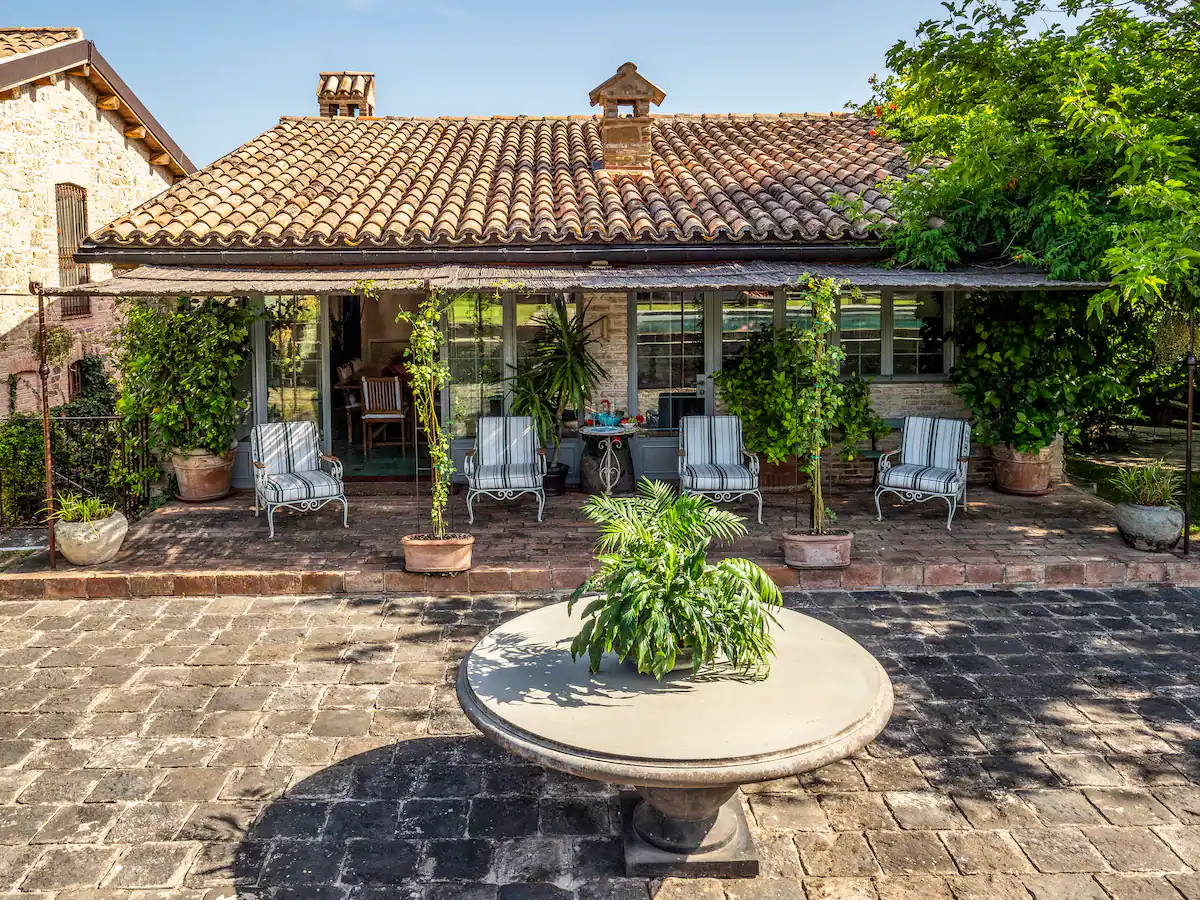 Casa con tetto di tegole, veranda con sedie a strisce, piante in vaso e tavolo rotondo in pietra su pavimento di mattoni.