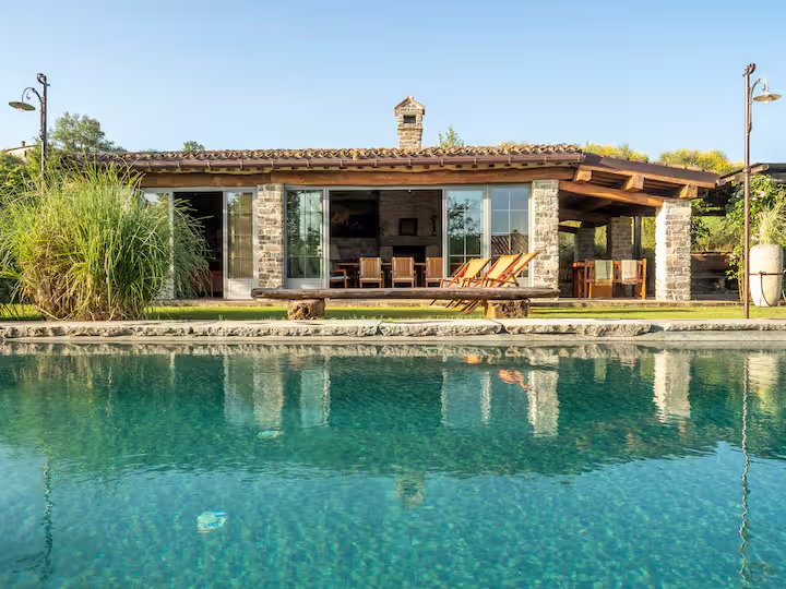 Villa con piscina e grandi vetrate, circondata da vegetazione, in una giornata soleggiata.