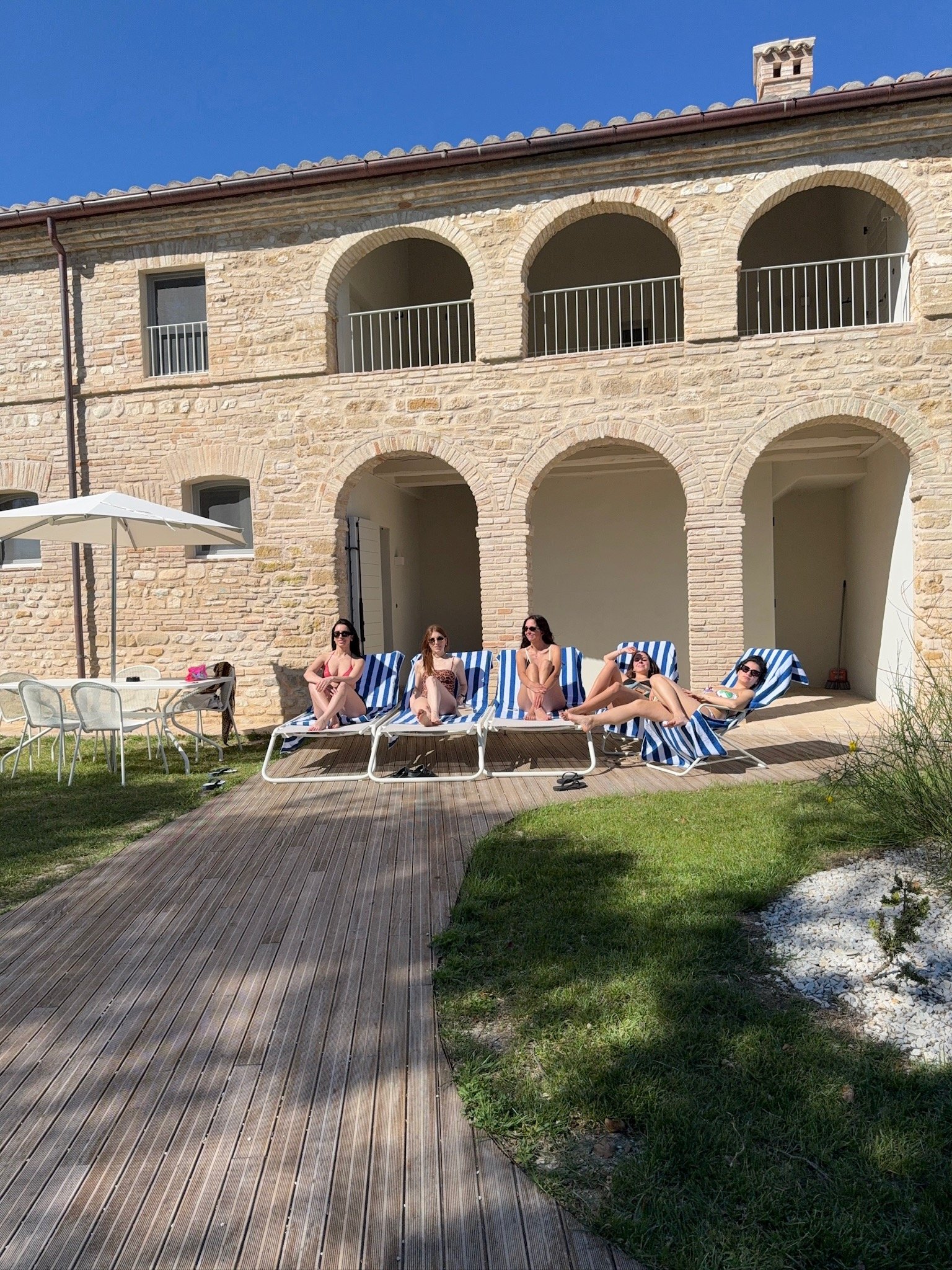 Quattro persone sdraiate su lettini a strisce blu e bianche davanti a un edificio di pietra con archi, su una terrazza di legno e erba.