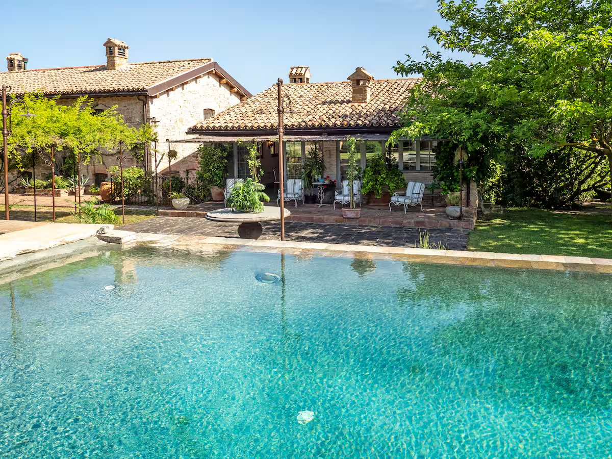 Una casa di campagna con piscina, circondata da vegetazione e dotata di una terrazza con sedie e piante.