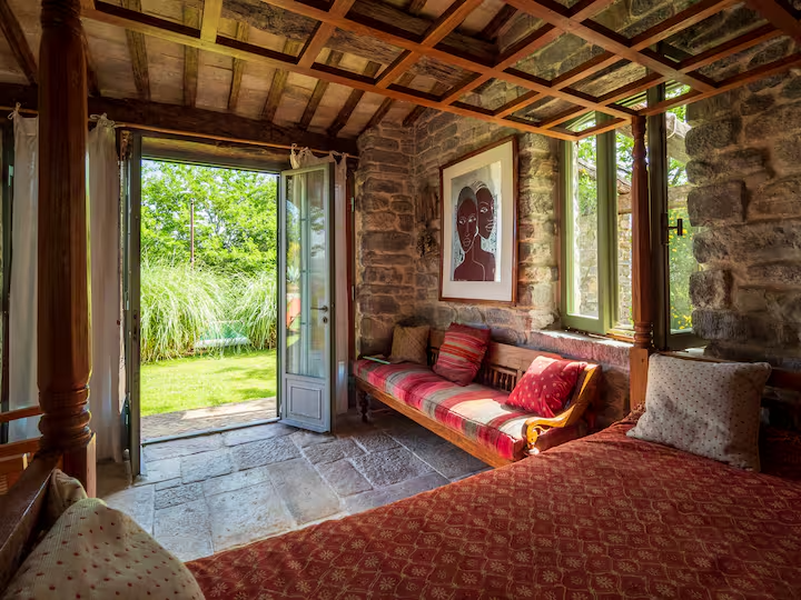 Interno di una stanza con pareti in pietra, un divano con cuscini rossi, un letto con copriletto rosso, una porta aperta verso un giardino verde e un quadro al muro.