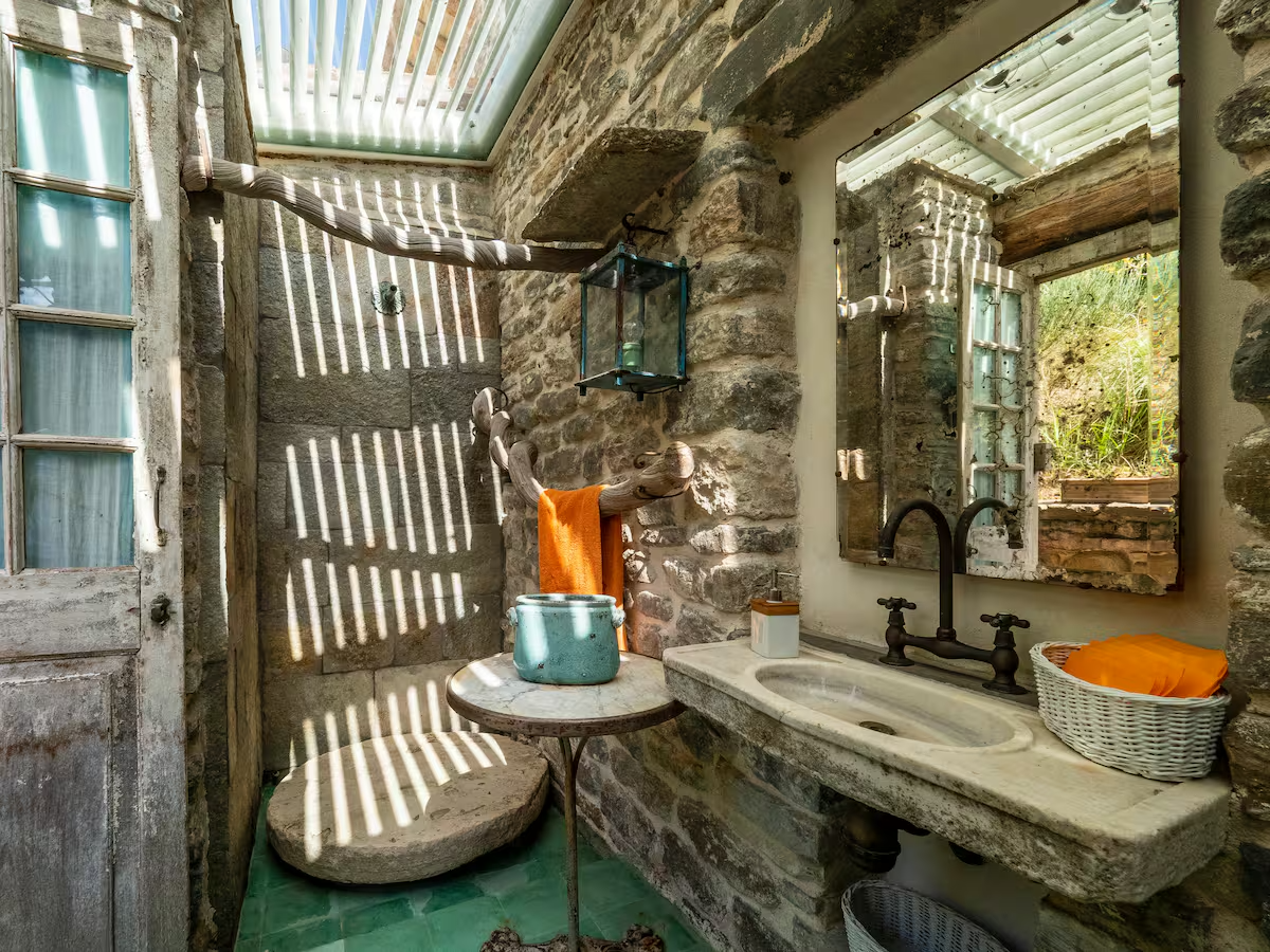 Bagno rustico con lavabo in pietra, specchio, pareti in pietra, tavolino con pianta e asciugamani arancioni, illuminato da luce naturale attraverso tetto trasparente.