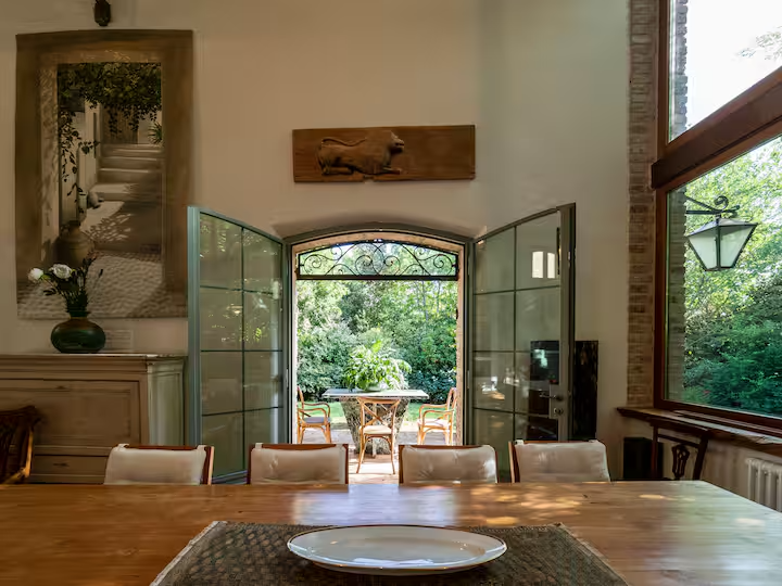 Sala da pranzo con tavolo in legno e sedie, porta aperta verso il giardino, grande finestra, decorazioni e veduta di alberi esterni.