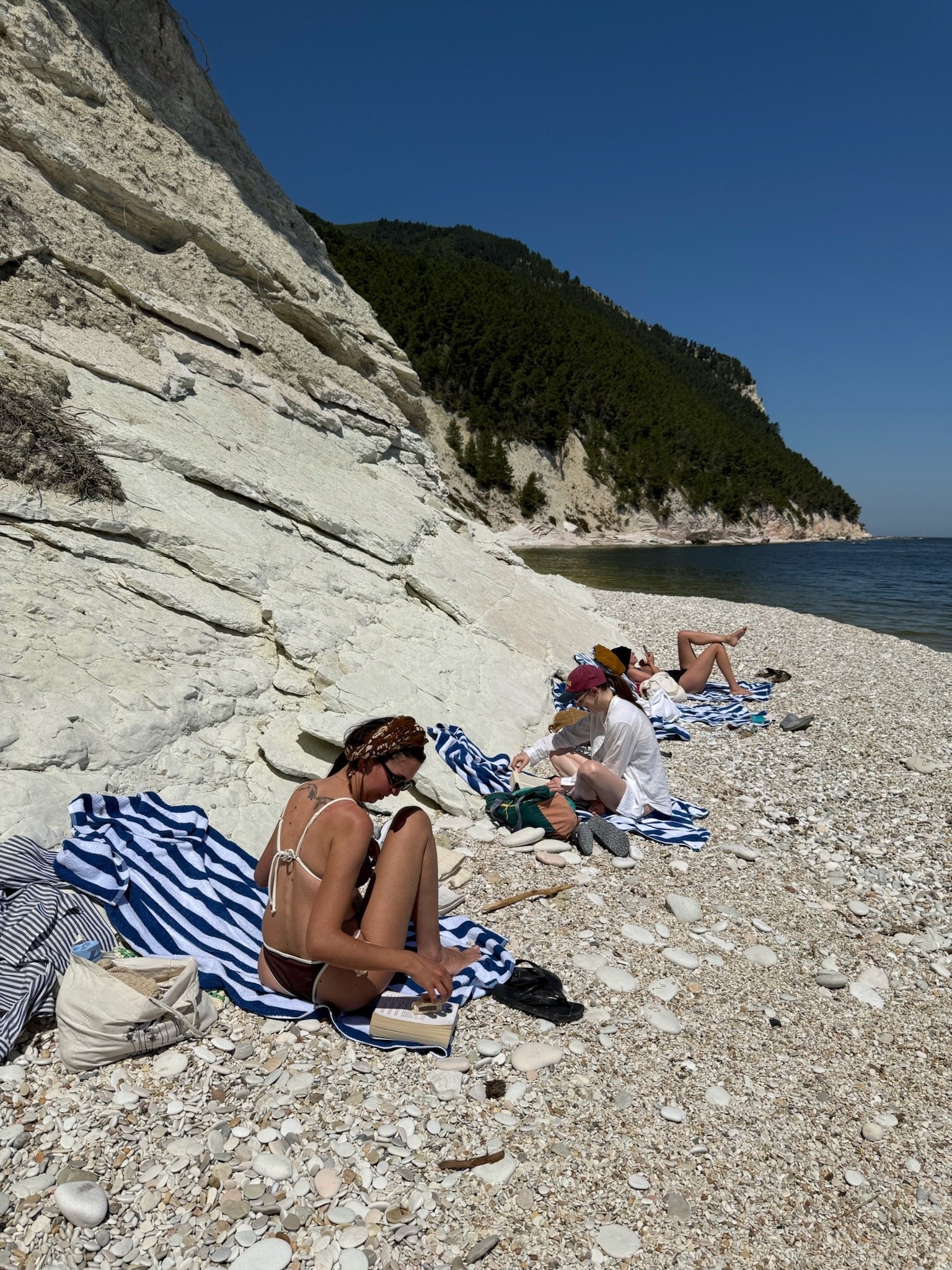 Gruppo di persone su una spiaggia di ciottoli, sdraiate su asciugamani a righe, con scogliere bianche e mare sullo sfondo.