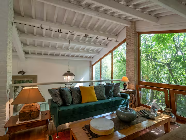 Soggiorno luminoso con soffitto in legno bianco, grandi finestre che mostrano alberi all'esterno, divano verde con cuscini colorati e tavolini in legno, lampade e accessori decorativi.