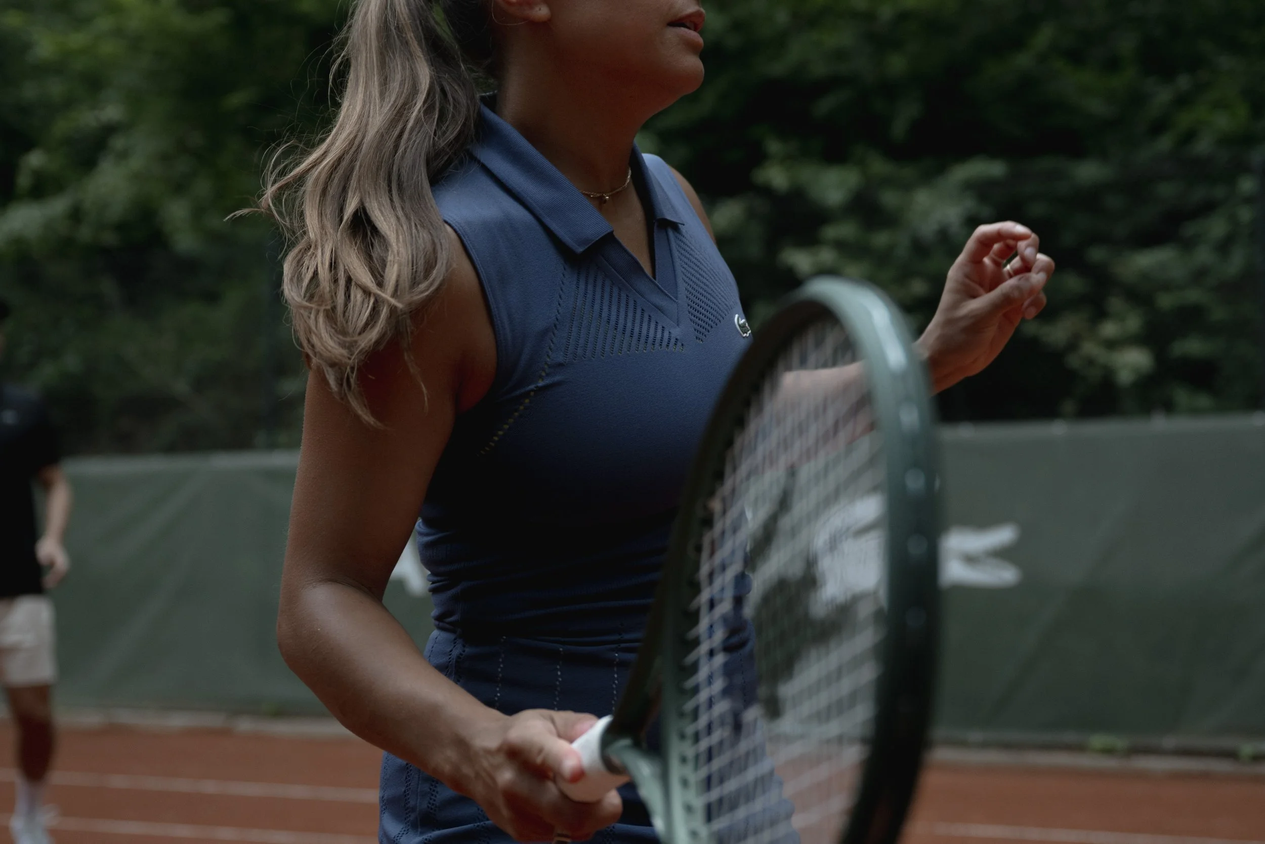 Person steht neben einem langen Tisch auf einem Tennisplatz, zum Essen vorbereitet, mit Lichterketten dekoriert. Im Hintergrund Bäume und blauer Himmel.