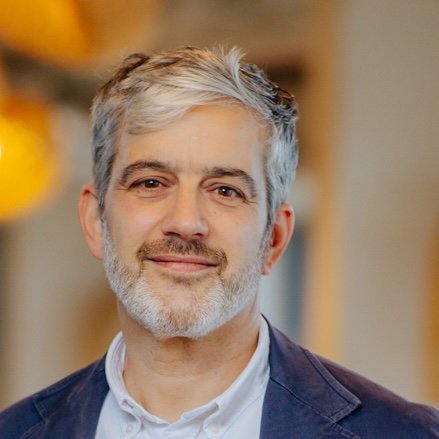 Smiling man with gray hair and beard wearing a jacket and white shirt indoors.