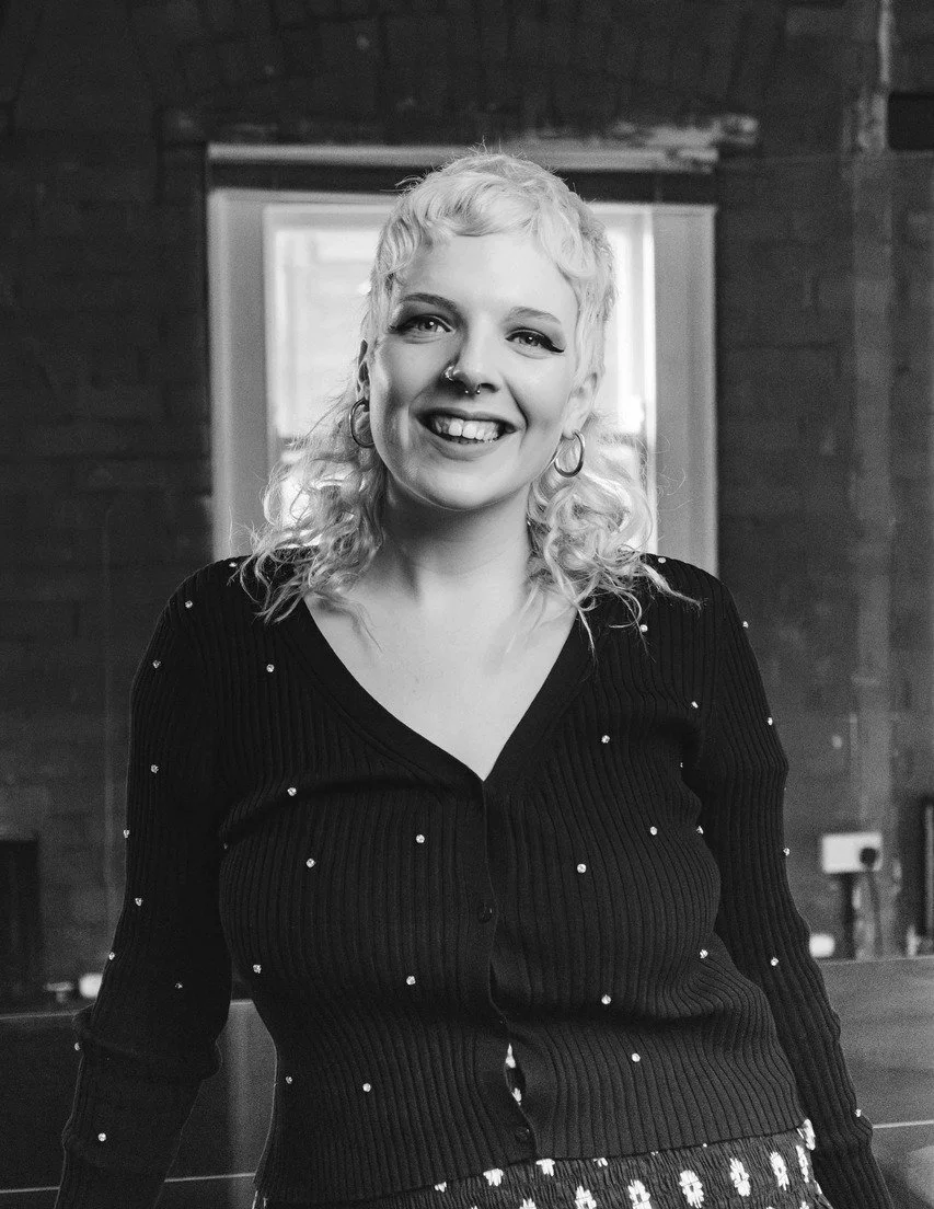 Julie Dunne stands in front of a brick wall. She is smiling directly at the camera and wearing a black cardigan.
