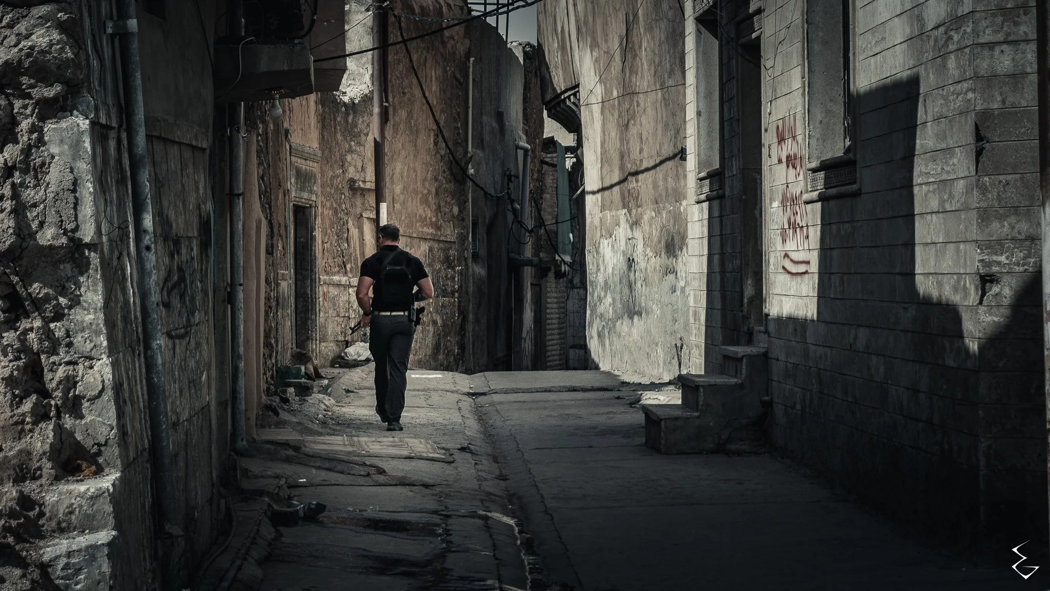 A man walking through a narrow, deserted alleyway with weathered and damaged walls on either side, showing signs of graffiti and decay, with shadows cast by surrounding buildings.