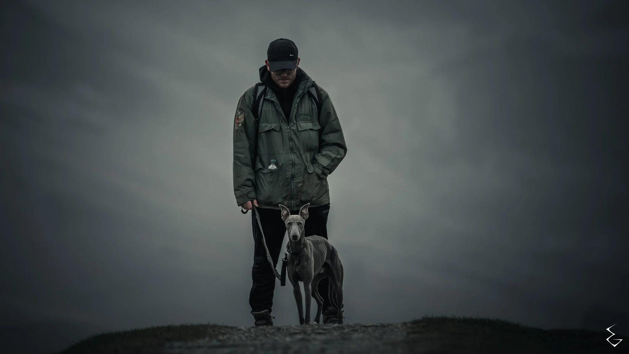 A man wearing a black cap, glasses, and a green jacket walking a grey dog on a leash during overcast weather.