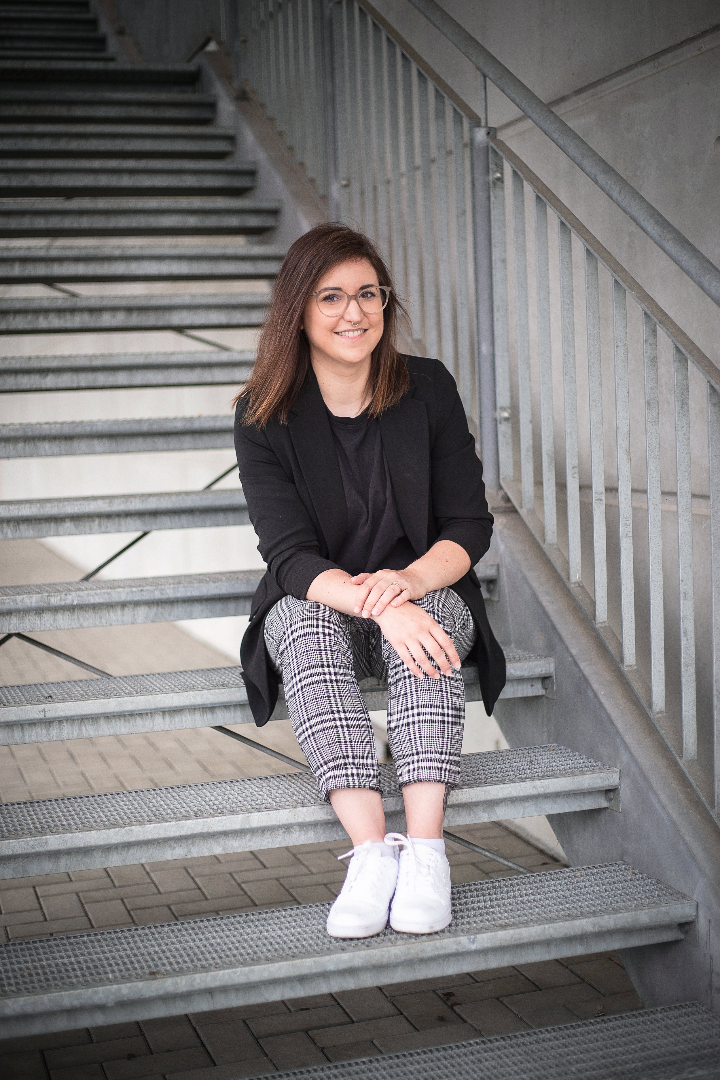Junge Frau mit Brille sitzt auf einer Industriestrumtreppe, trägt eine schwarze Jacke, karierte Hosen und weiße Sneakers.
