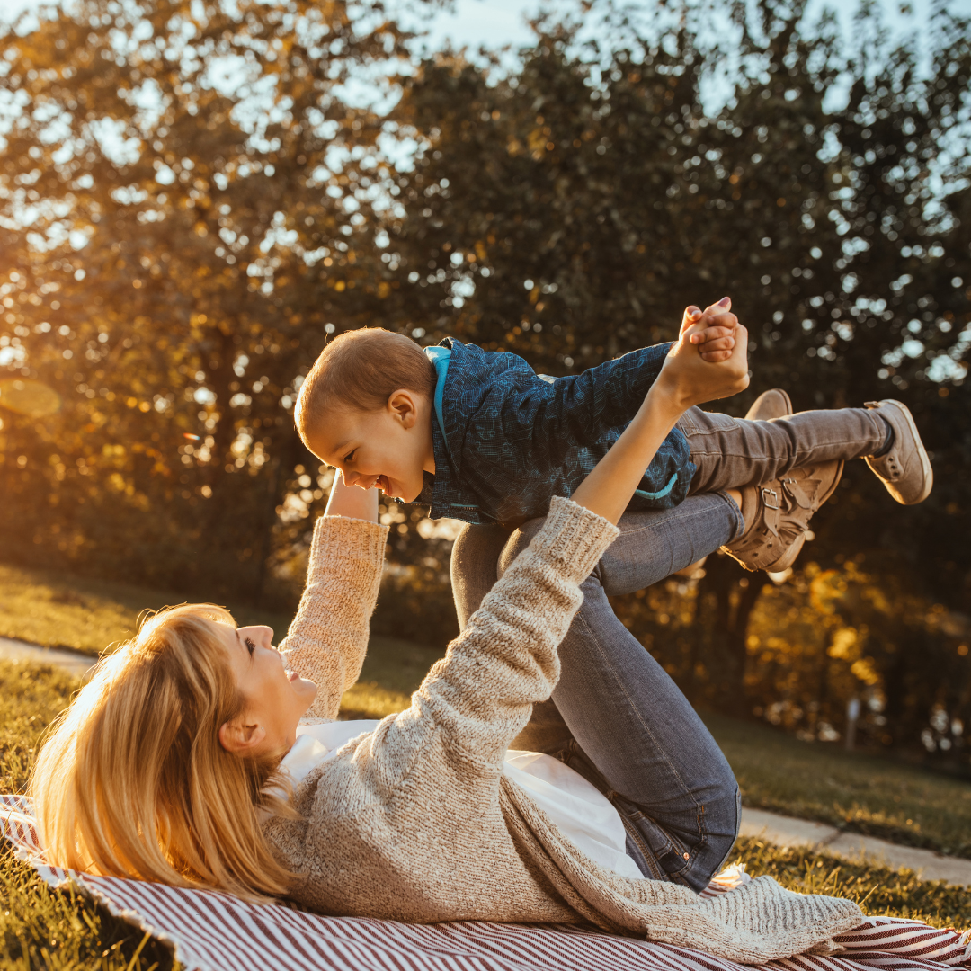 A woman and a young boy play together outdoors on a blanket during sunset. The woman is lying on her back on the blanket, lifting the boy in the air with her hands. Both are smiling and enjoying the moment amid trees with orange leaves.