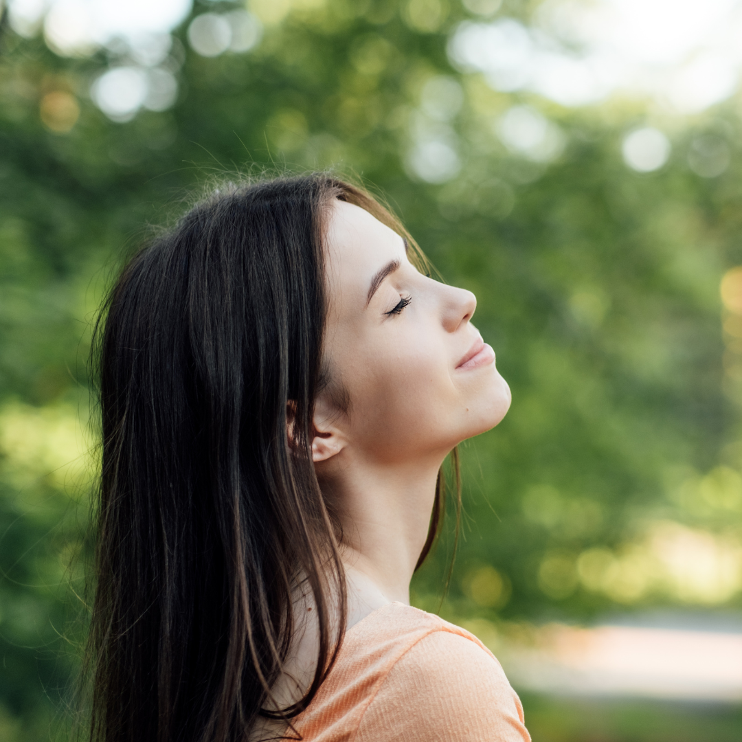 Young woman with long dark hair smiling and relaxing outdoors with eyes closed, surrounded by greenery.