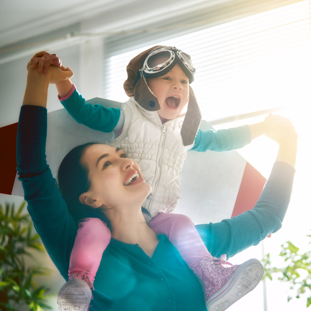 A woman lifting her young daughter, who is wearing aviator goggles and a pilot's hat, high in the air indoors with sunlight coming through the window.
