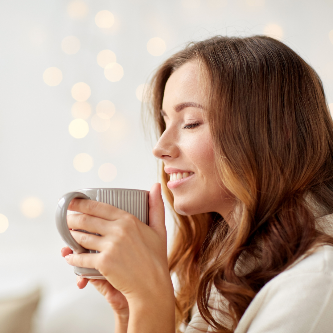 A woman with red hair smiling and holding a gray mug with both hands.