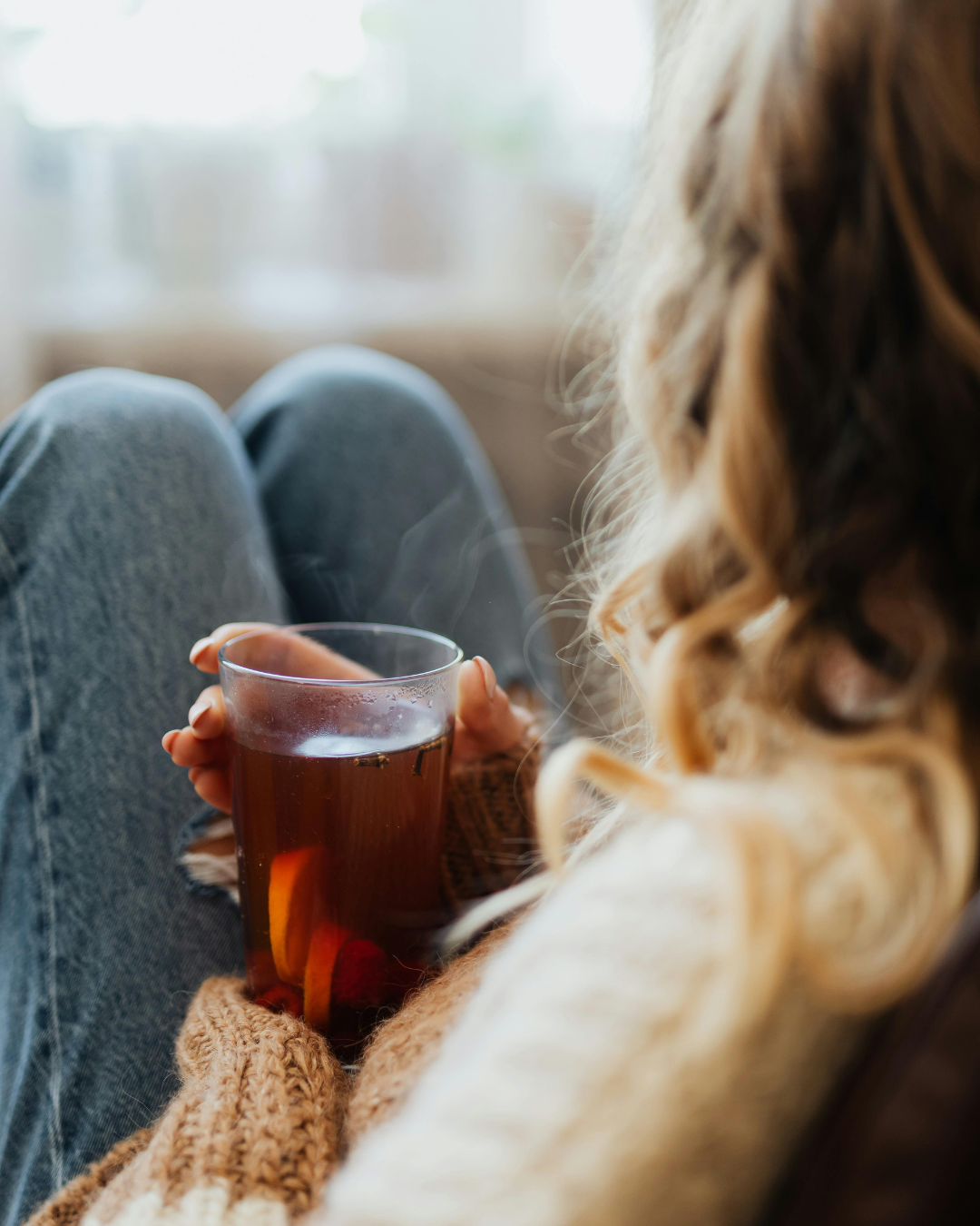 A woman holding a glass mug of tea or coffee, sitting comfortably with her knees up, in a cozy indoor setting with soft natural light.