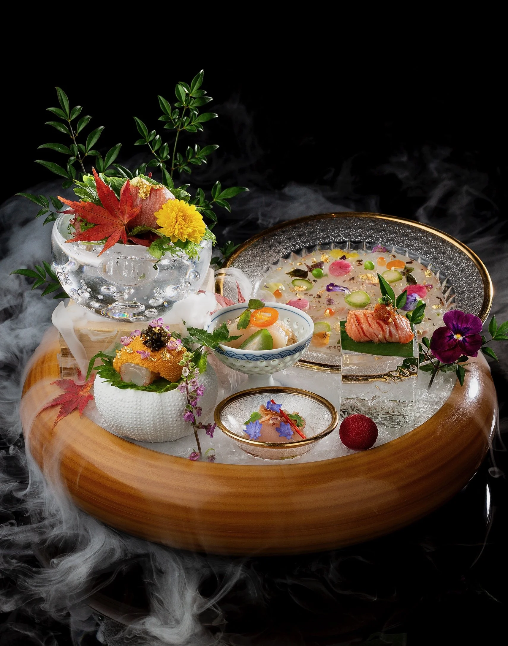 Assorted traditional Japanese dishes arranged on a circular wooden tray with layered glass plates, garnished with flowers, leaves, and decorative elements, with mist or smoke surrounding them.