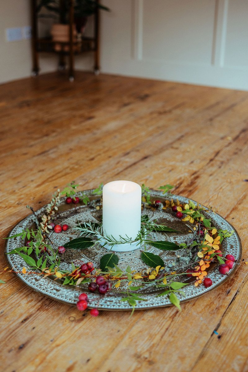 A white pillar candle on a decorative metallic tray surrounded by green leaves, red berries, and yellow and orange foliage on a wooden table.