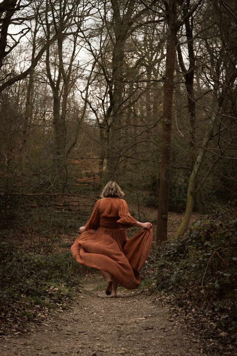 A woman in a long, flowing dress walking through a wooded area on a dirt path.