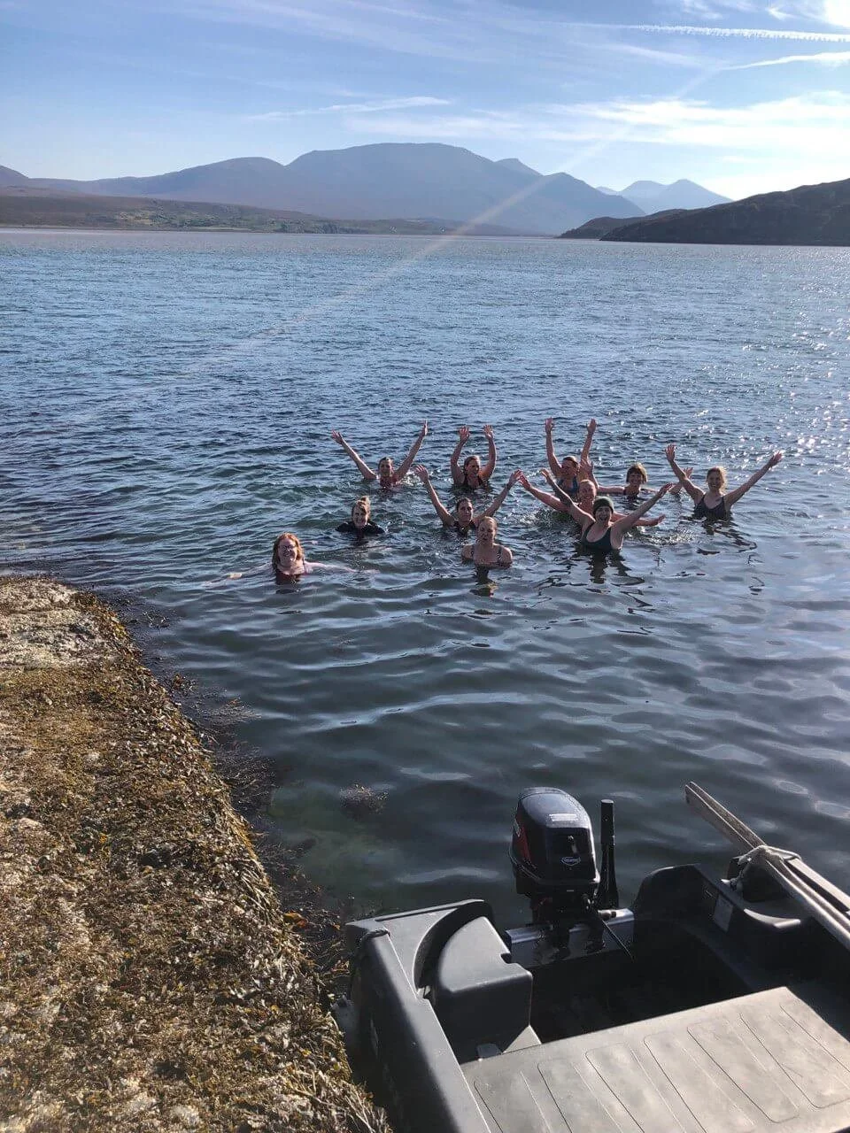 A group of people swimming in a lake near a rocky shore with mountains in the background under a blue sky.