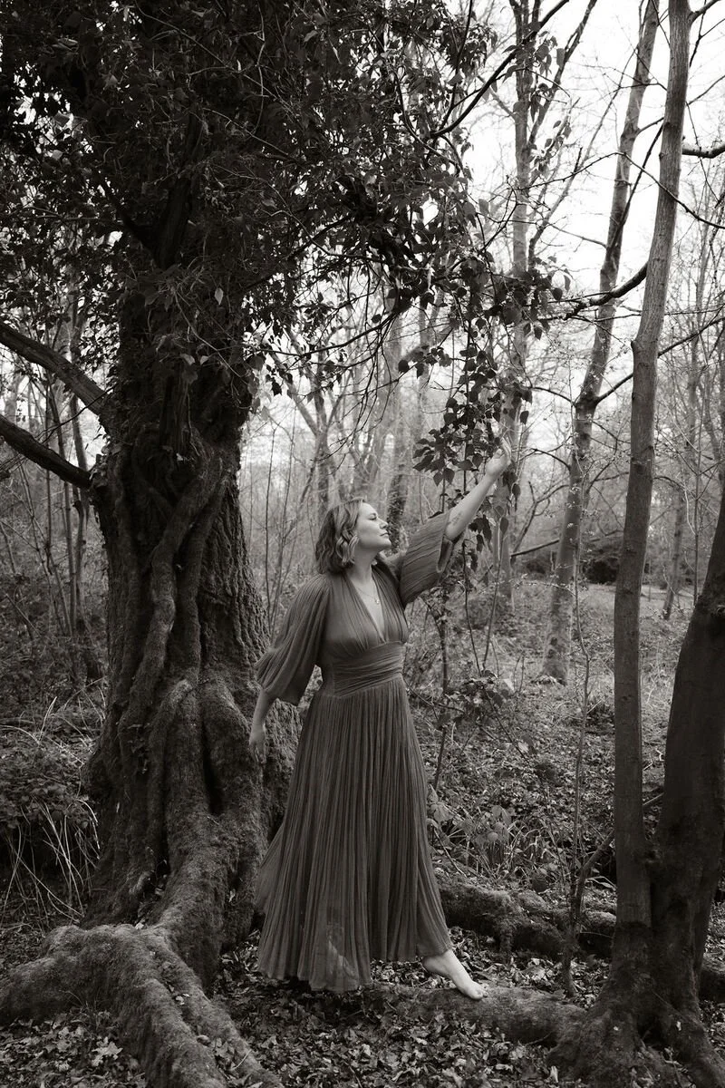 A woman in a long pleated dress standing barefoot outdoors in a forest, reaching up and touching the branches of a tree.