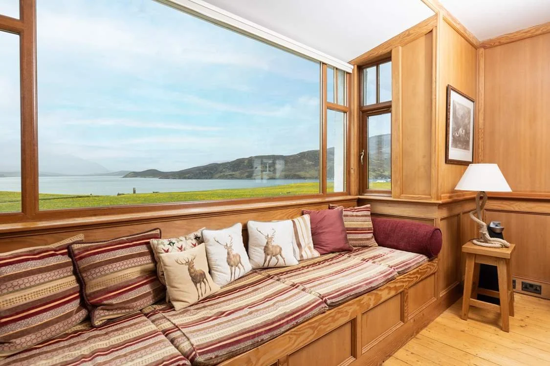 Living room with large window view of hills and water, wooden walls, a cushioned window seat with pillows featuring deer, and a side table with a lamp.