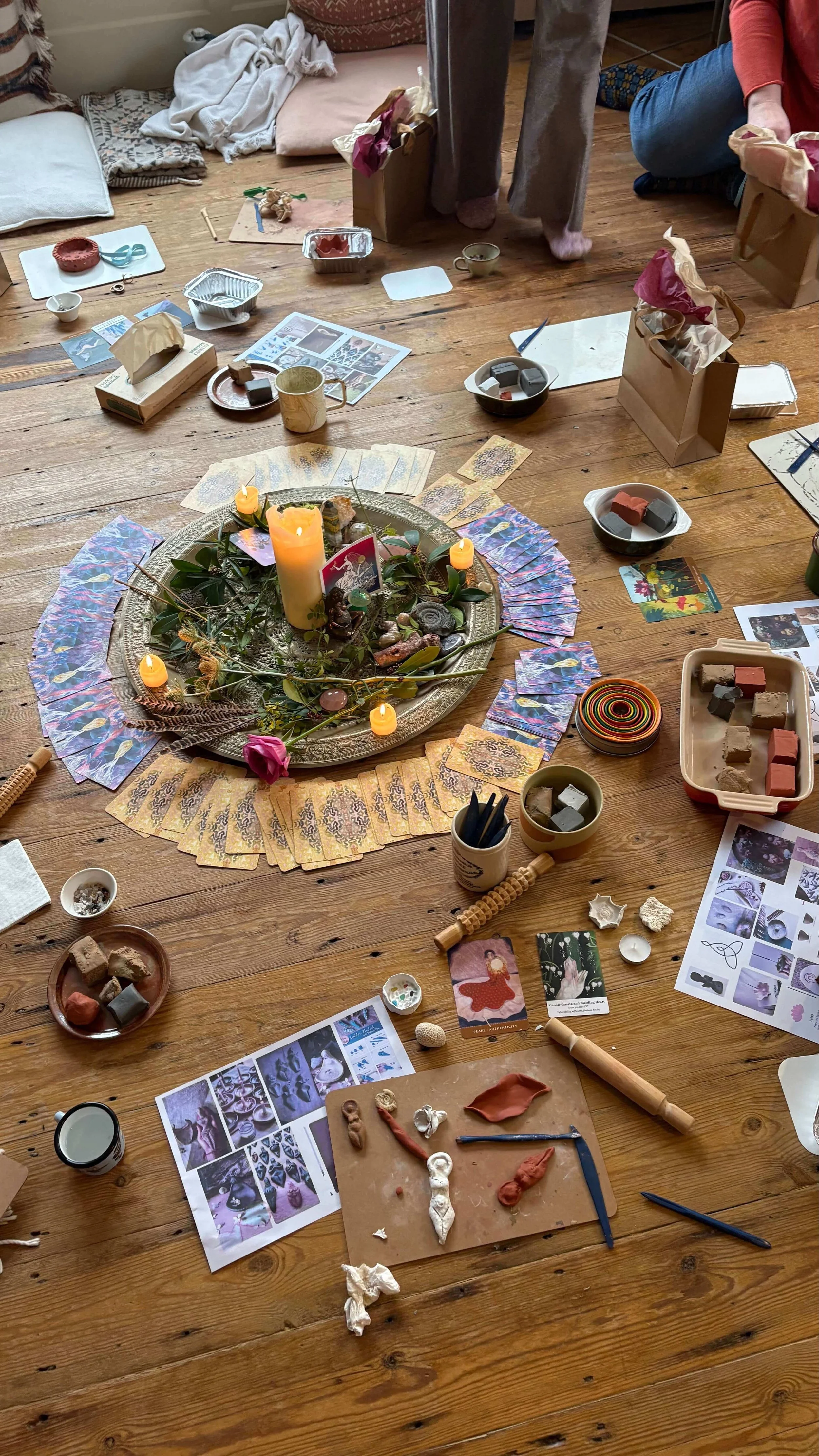 Table with candles, flowers, seashells, cards with affirmations, a basket of paint tubes, a plate with paint, a glass of red liquid, and notecards.