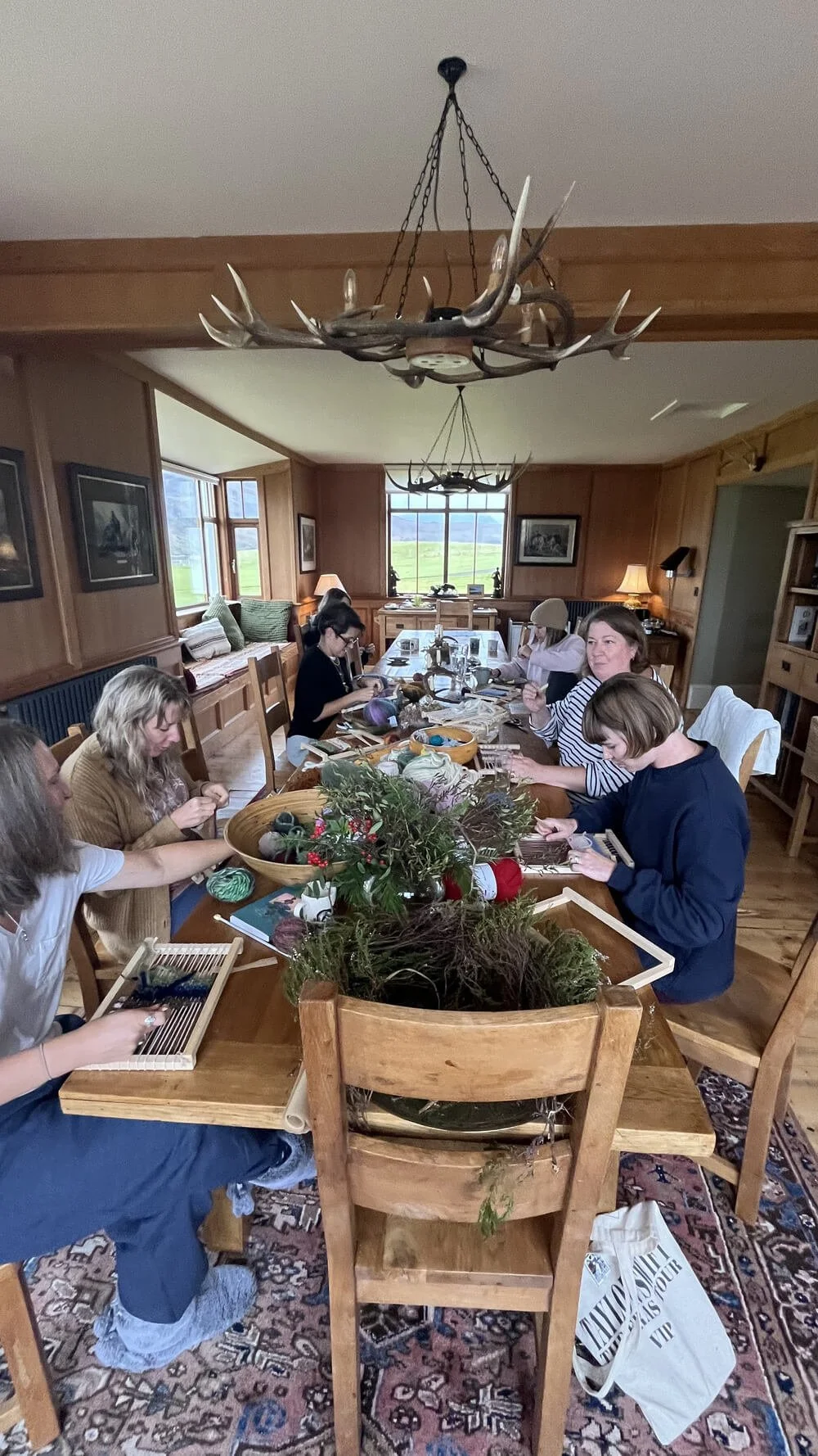 A group of people sitting around a wooden dining table engaged in a craft activity, with holiday decorations and natural light from windows in a cozy, wood-paneled room.