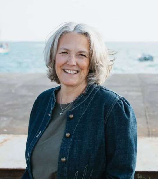 Smiling woman with shoulder-length grey hair standing outdoors near a body of water with boats in the background.