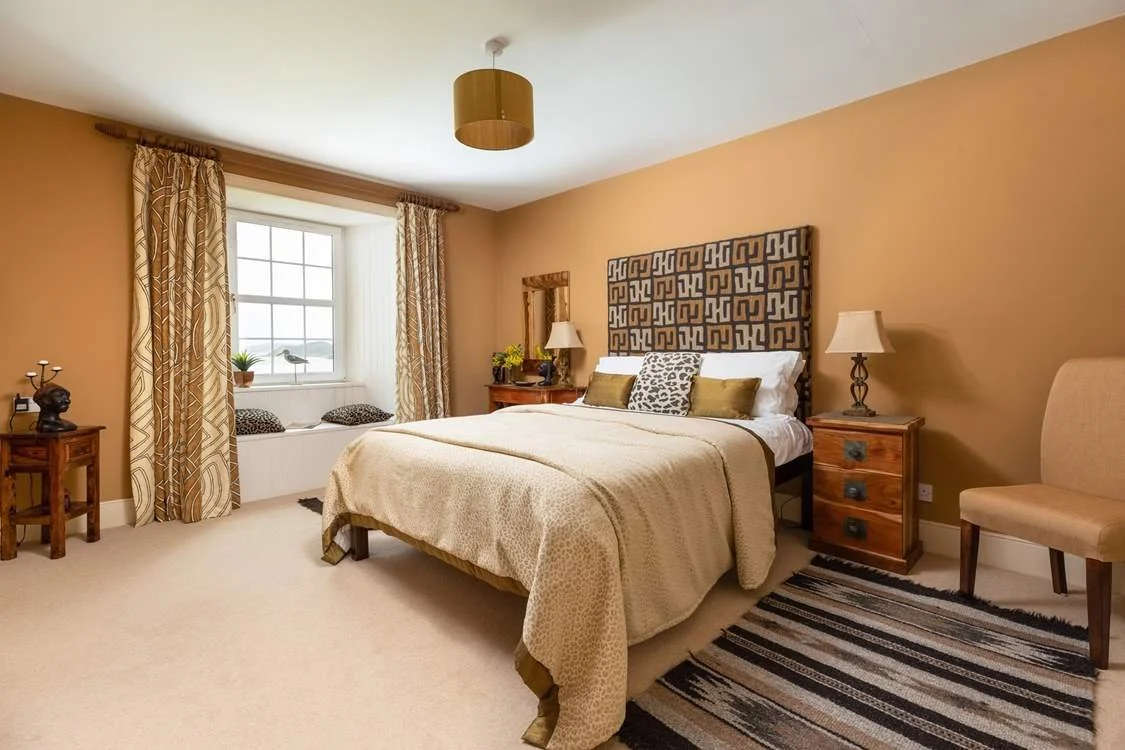 A cosy bedroom with a large bed, decorative headboard, two nightstands with lamps, a window seat with cushions, and a beige chair, decorated in warm tones.