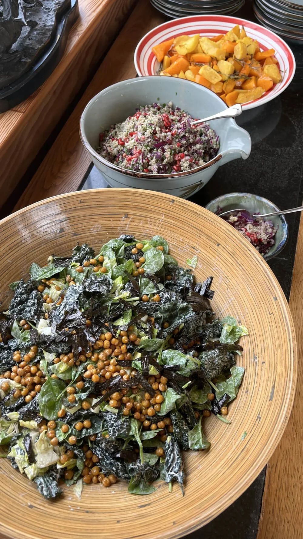 Mixed salad with dark leafy greens and chickpeas in a large wooden bowl, with other bowls of colorful chopped vegetables and shredded cheese nearby.