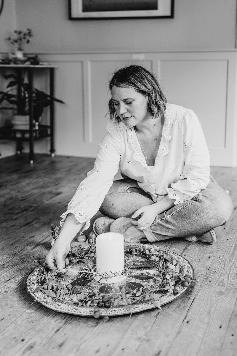 A woman sits on the floor arranging a decorative wreath with a white candle in the center on a patterned tray.