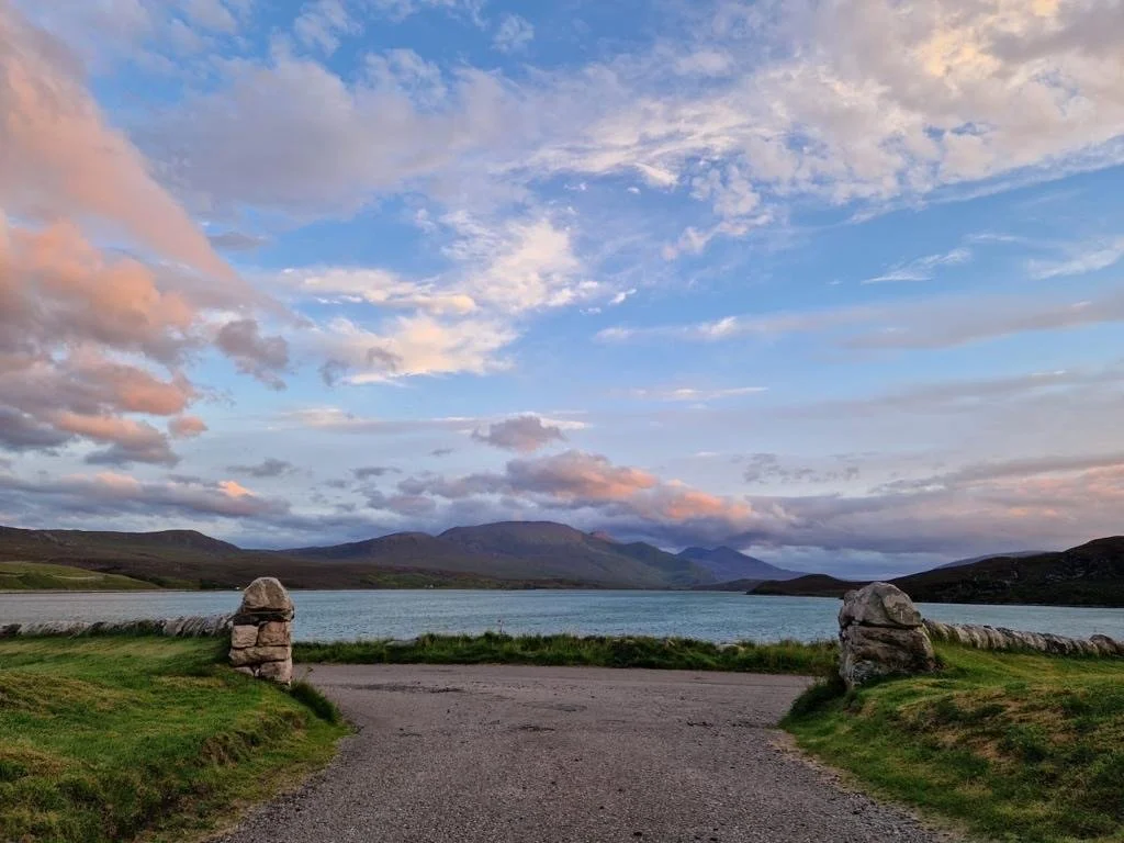 Scenic view of a large lake surrounded by green grassy areas, stone markers on either side of a gravel path, and distant mountains under a partly cloudy sky during sunset.