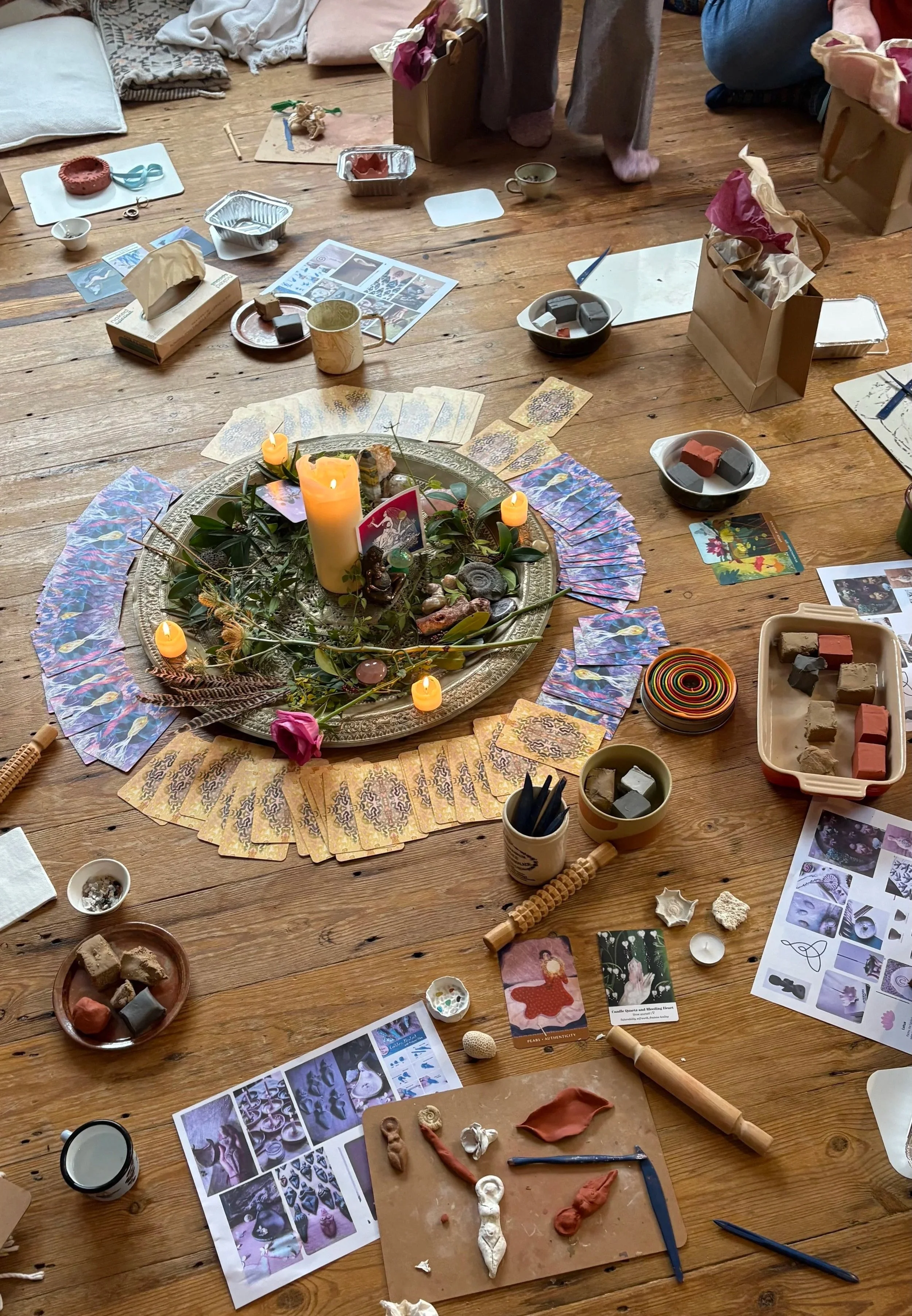 Table with candles, flowers, seashells, cards with affirmations, a basket of paint tubes, a plate with paint, a glass of red liquid, and notecards.