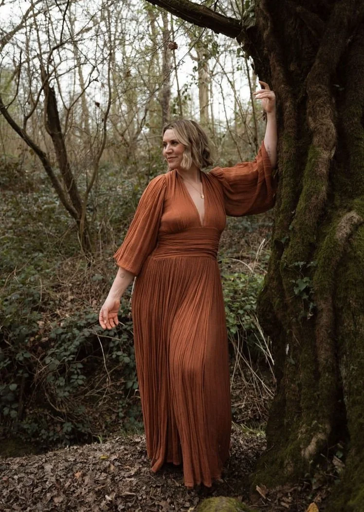 A woman with shoulder-length blonde hair wearing a rust-colored, pleated maxi dress with three-quarter sleeves stands outdoors next to a large tree with moss. She is smiling slightly and looking to her right in a wooded area with leafless trees and dense foliage.