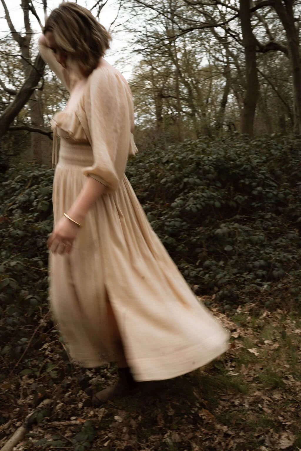 A woman in a beige dress walking through a wooded area with trees and foliage.