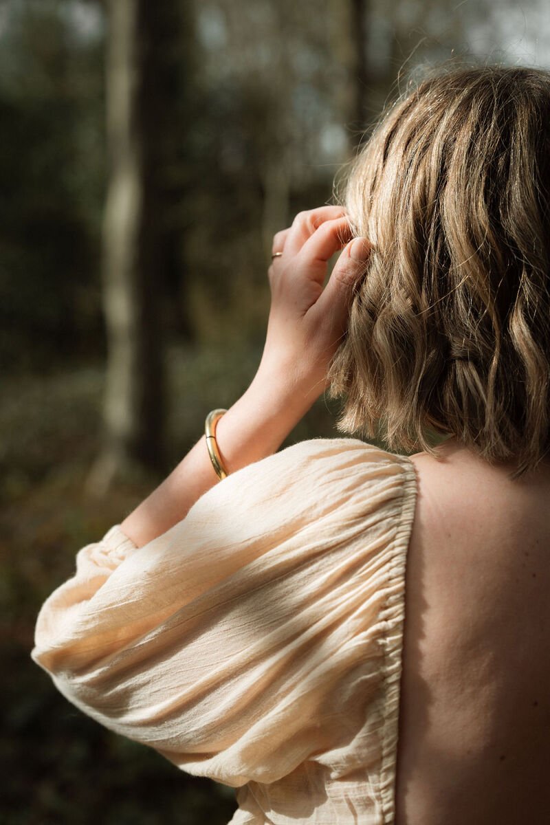 A woman with light brown, wavy hair wearing a beige puff-sleeve top, touching her ear in a forest setting with blurred trees in the background.