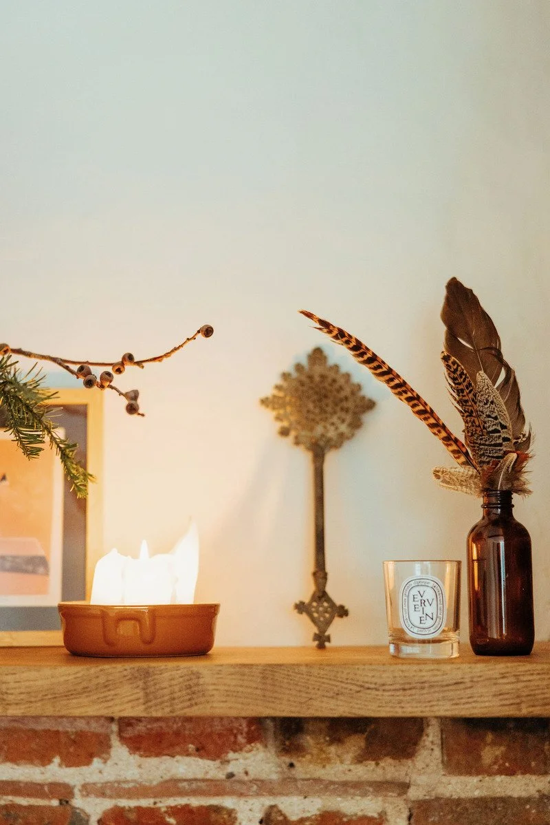Decorative display on a wooden surface with a burning candle in a ceramic dish, a glass with the word 'Evergreen', and a brown bottle holding feathers. A metal decorative item is mounted on the wall behind, with a small branch with berries on the left side.