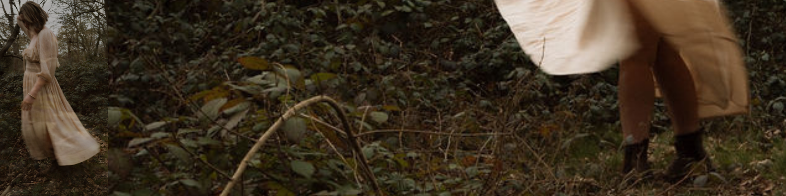 A person wearing a long beige dress and black boots standing and bending over in a wooded area with plentiful greenery.