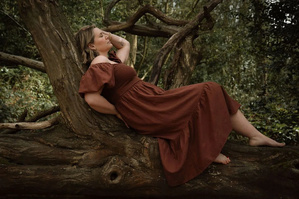 A woman in a brown, off-shoulder dress reclining against a tree in a forest setting.