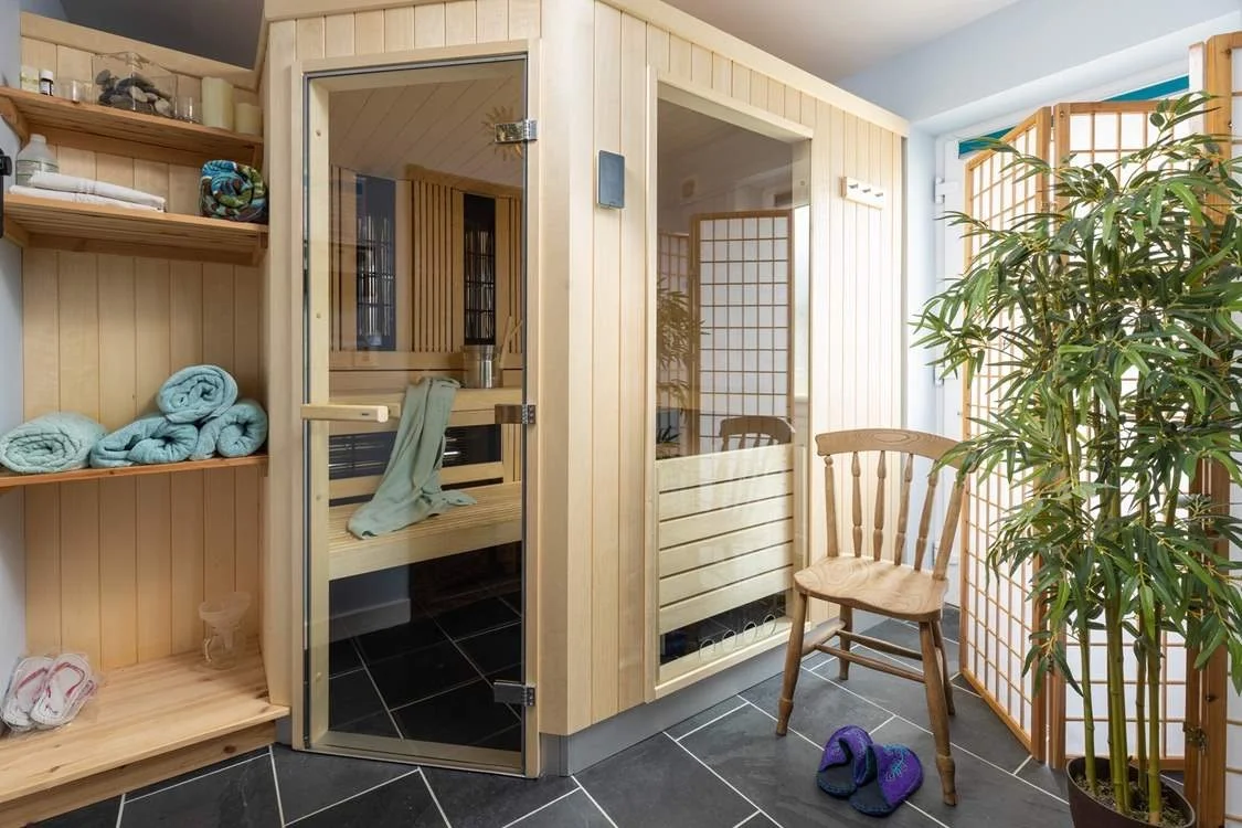 Interior of a sauna room with glass doors, wooden shelving with rolled towels, a wooden chair, and a large potted plant.