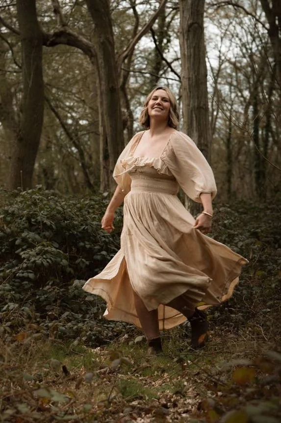 A woman in a flowing, beige dress smiling and twirling in a forest with bare trees and low shrubs.