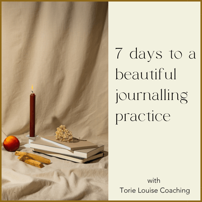 A red candle, an apple, and a few biscuits sit on a cloth next to a stack of books with a floral decoration, with a sign that reads '7 days to a beautiful journaling practice with Torie Louise Coaching'.