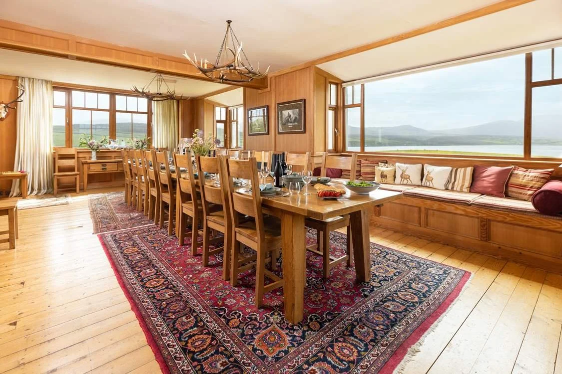 Dining room with wooden furniture, chandeliers, large windows showing an outdoor view, and decorated with rugs and pillows.