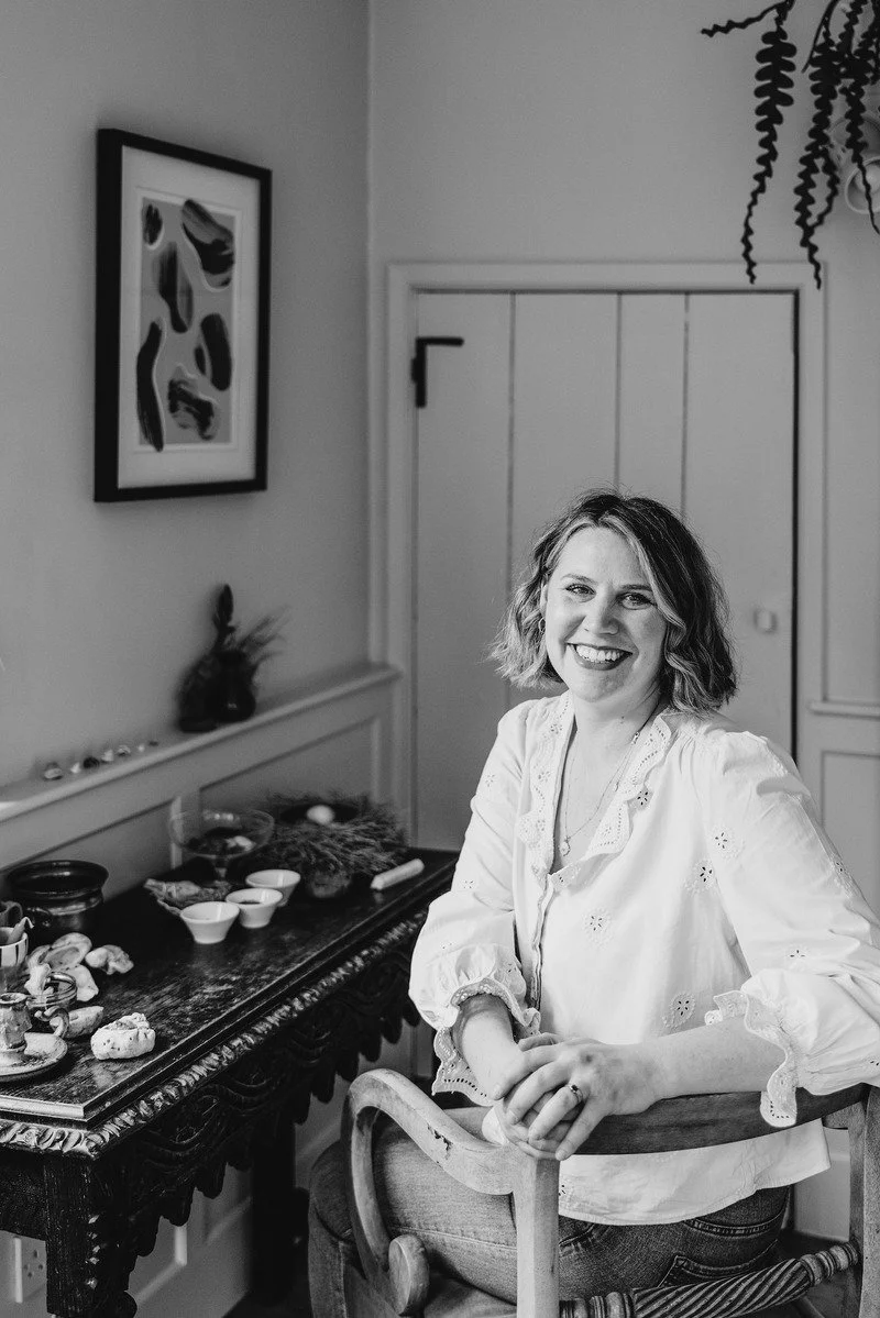 A woman with shoulder-length hair smiling and sitting on a wooden chair in a room with a decorated table beside her, on which there are various bowls and objects. The room has a framed abstract art piece on the wall and a door in the background.