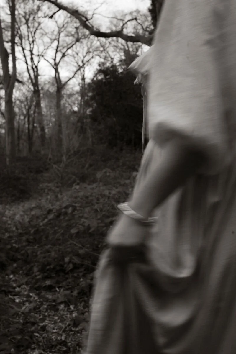 A blurred person wearing a light-colored jacket standing in a wooded outdoor area with leafless trees and ground covered in leaves.