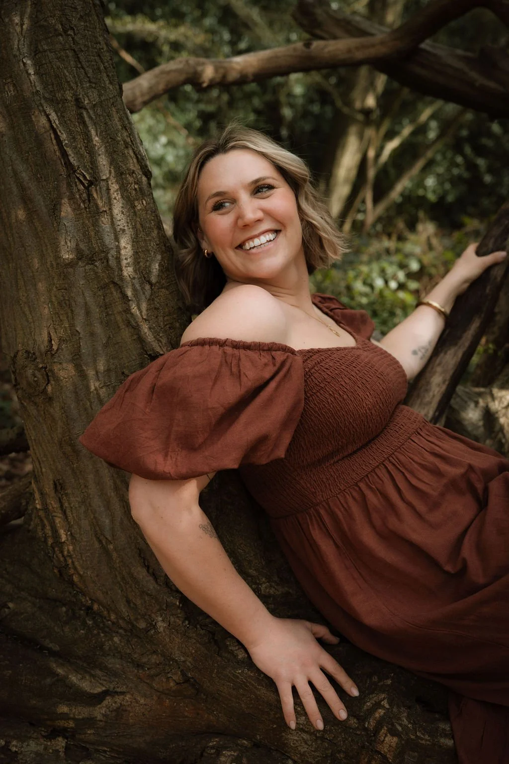 A woman with short blonde hair and a big smile, wearing an off-shoulder brown dress, leaning against a tree in a forested area.