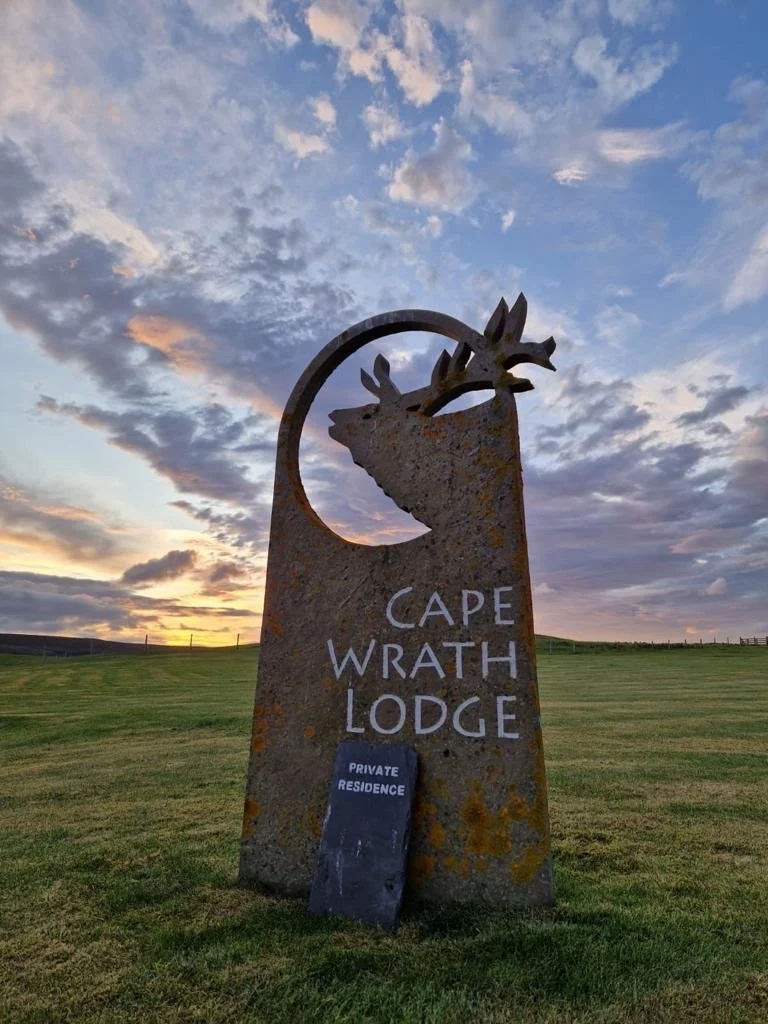 A large rusted stone sign for Cape Wrath Lodge with a relief of a stag's head at the top, set on a grassy landscape during sunset with a dramatic cloudy sky. A smaller black sign reading 'Private Residence' leans against it.