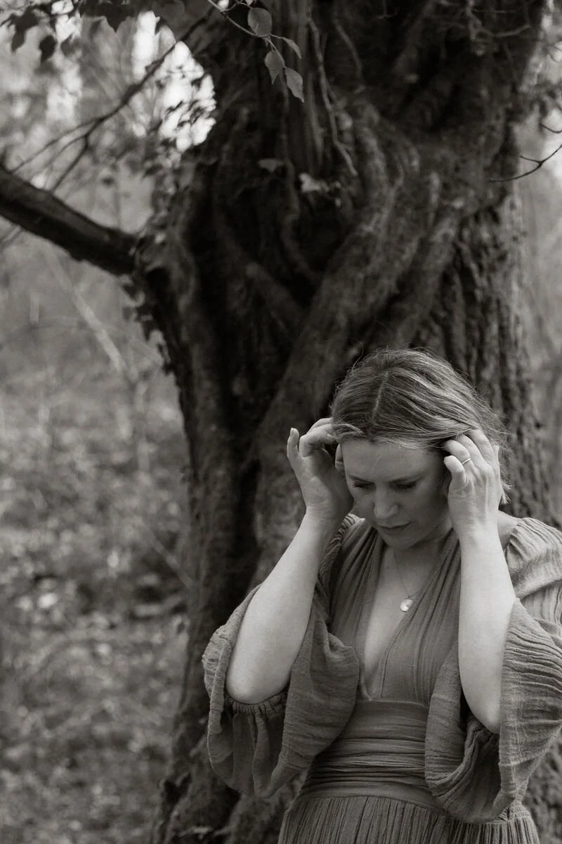 A woman with short hair standing outdoors in front of a large tree, touching her head with both hands, appearing thoughtful or concerned.